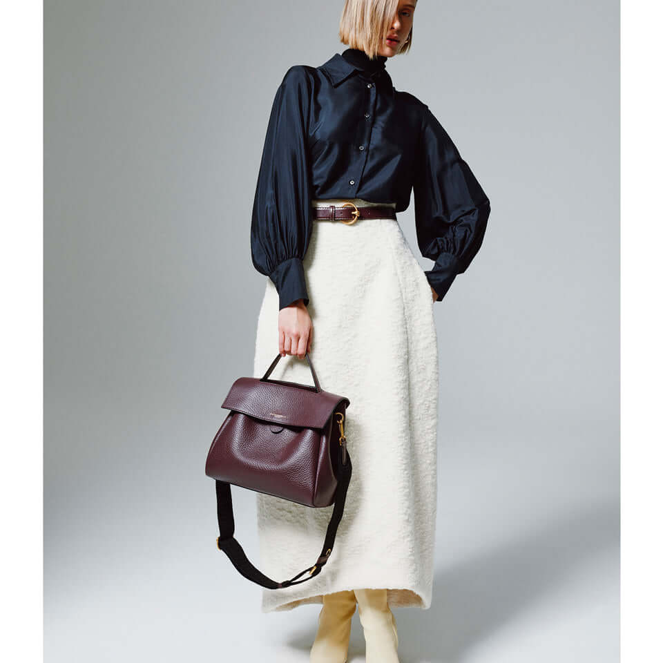 Model wearing a navy blouse and white skirt, holding the Clio handbag by Gianni Chiarini in burgundy.