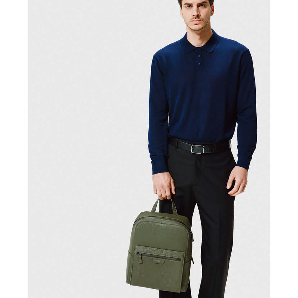Man in navy polo and black pants holding a green backpack, showcasing a modern and functional accessory for men.