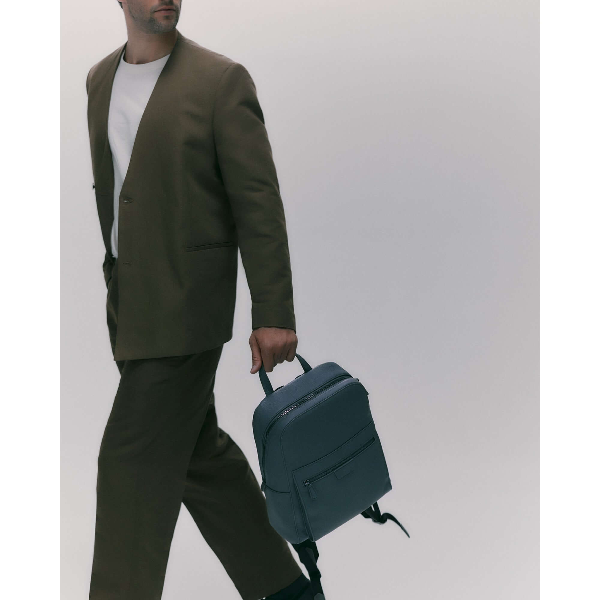 Man carrying a stylish blue leather backpack while wearing a tailored brown suit.